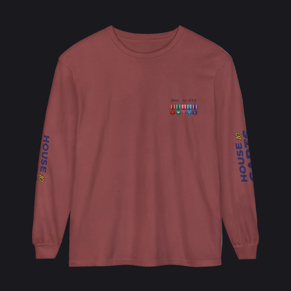 A long-sleeve crewneck t-shirt in a dusty rose or mauve color, featuring a small military-style ribbon graphic on the left chest, 'HOUSE 98' printed in blue on both sleeves, and a small yellow crown detail on the right sleeve cuff.