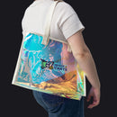 A person wearing a white t-shirt and blue jeans carries a large, semi-transparent holographic tote bag with cream-colored shoulder straps. The bag features a colorful iridescent print with a 'House of Carts' logo printed on the front.