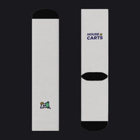 A pair of white crew socks shown from both the front and back views against a plain white background. The front view features a small multicolor logo near the foot, while the back view features the 'HOUSE OF CARTS' logo in navy blue and gold near the calf, with a black ribbed heel and toe.