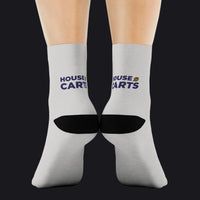 A pair of crew socks worn on feet, shown from the back. The socks are light gray with a black heel section and navy blue text reading 'HOUSE OF CARTS' with a small yellow graphic accent.