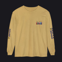 A long-sleeve crewneck t-shirt in a sandy tan color, featuring a small multicolor military-style ribbon graphic on the left chest with the text 'MWK 3RTO' above it. The right sleeve has 'HOUSE CARDS' printed vertically in dark blue, and the left sleeve has a matching logo on the opposite side.