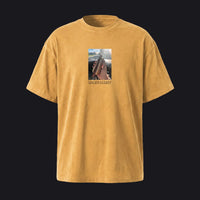 A mustard yellow short-sleeve t-shirt with a central graphic print showing a hand holding a rifle inside a car, with the text 'LUCKY GLZZY' printed below the image, displayed against a solid light gray background.
